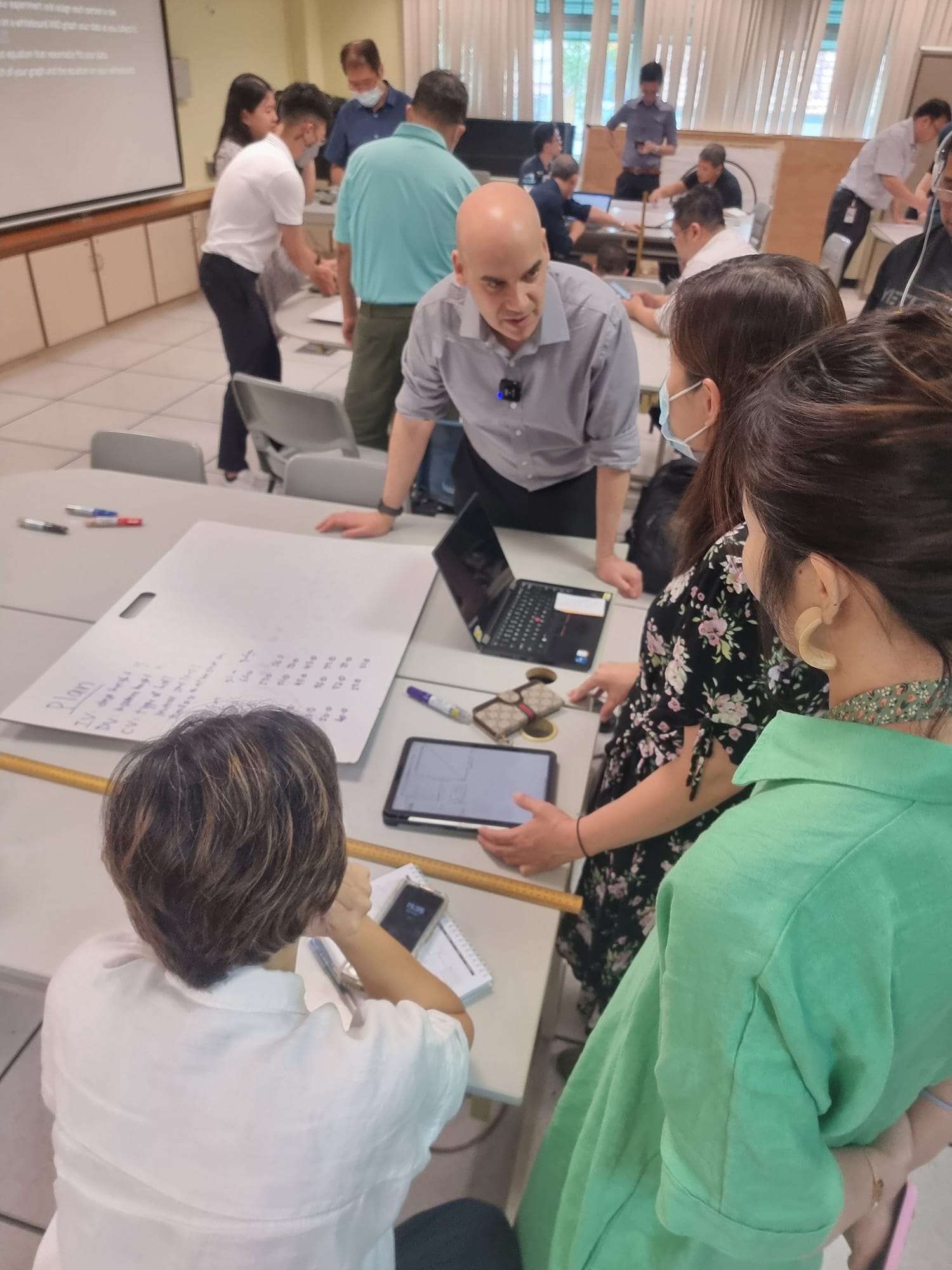 Several people gathered around a table in a workshop room, discussing over a tablet, laptop, and written notes.